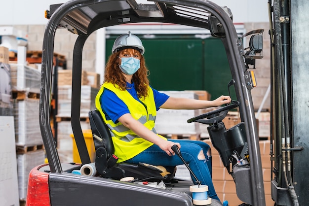 An industrial female worker wearing a medical mask using a forklift in a warehouse