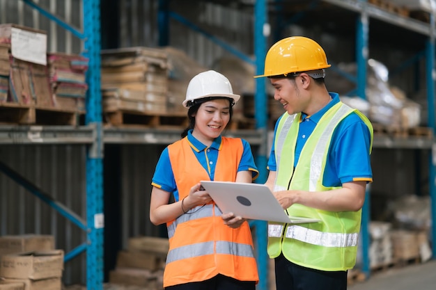 Industrial warehouse asian worker man and woman talking smiling and looking laptop and using barcode scanner checking goods and box shelf stock in warehouse factory store Warehouse Logistic concept