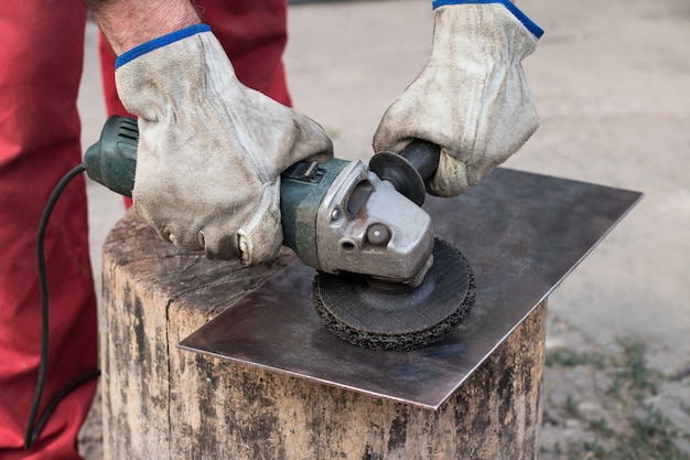 An industrial worker in a red overall with his hands in working gloves grinds a piece of sheet steel with an old grinder