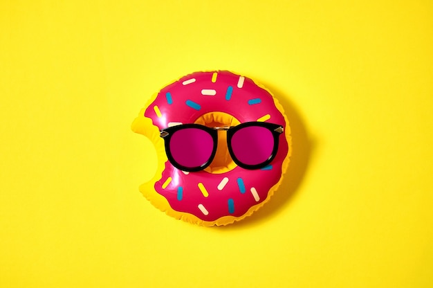 Inflatable donut in pink sunglasses on yellow background. Creative minimal concept. Top view Flat lay