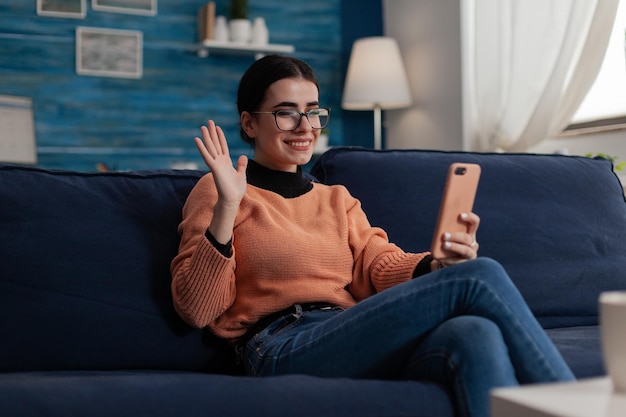 Influencer on couch holding smartphone waves hand at camera. Smiling student with glasses holding mobile phone on sofa in remote videocall. Digital content creator posting on social media.