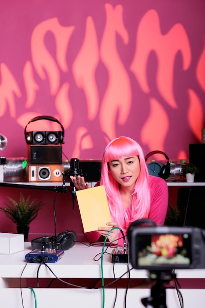 Influencer with pink hair presenting notebook in front of camera recording school supplies review using professional vlogging equipment. Asian vlogger filming podcast, live streaming