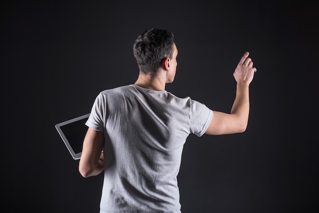 Information technology. Smart handsome IT specialist standing in front of the sensory screen and using it while having a laptop in his hand