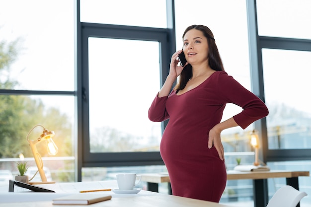 Informative talk. Waist up of charming pregnant woman having conversation on mobile phone while looking away and touching her back