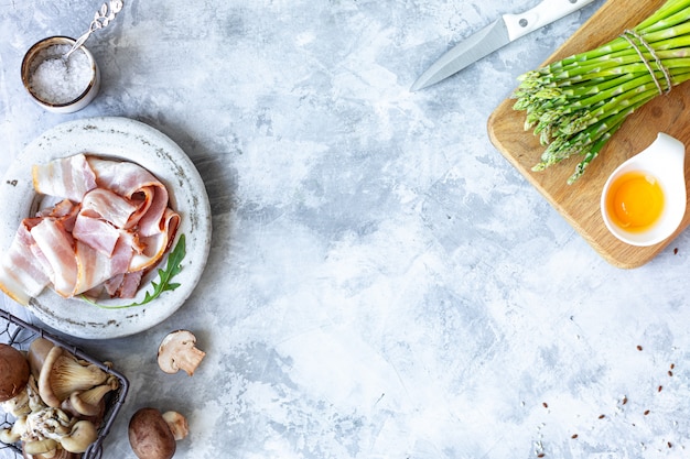 Ingredients for cooking on a gray concrete background. A bunch of fresh green asparagus, eggs, bacon, mushrooms. Top view. Copy space