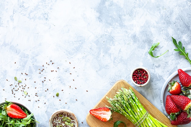 Ingredients for cooking on a gray concrete background. A bunch of fresh green asparagus, strawberries, arugula. Top view. Copy space