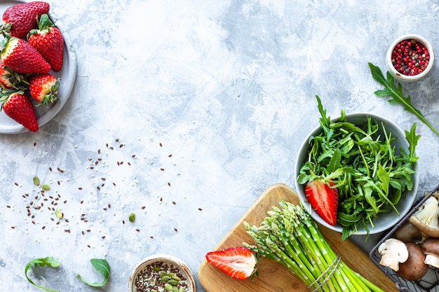 Ingredients for cooking on a gray concrete background. A bunch of fresh green asparagus, strawberries, mushrooms, arugula. Top view. Copy space