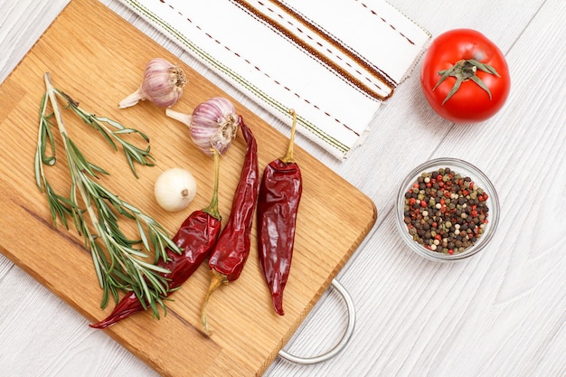 Ingredients for cooking meat or fish. Garlic, allspice peppers in glass bowl, chili pepper, tomato, onion and rosemary on a wooden cutting board and napkin. Top view.