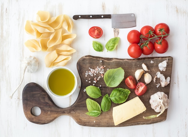 Ingredients for cooking pasta. Conchiglioni, basil leaves, cherry-tomatoes, Parmesan cheese, olive oil, salt, garlic on rustic walnut chopping board over white wooden 