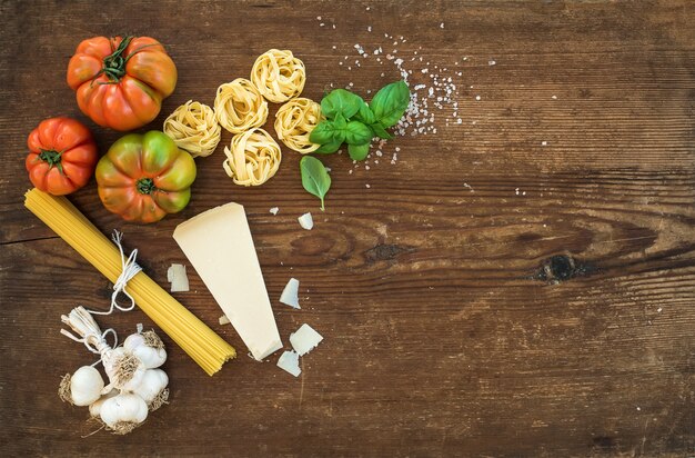 Ingredients for cooking pasta. Spaghetti, tagliatelle, garlic, Parmesan cheese, tomatoes and fresh basil on rustic wooden background, top view, copy space.