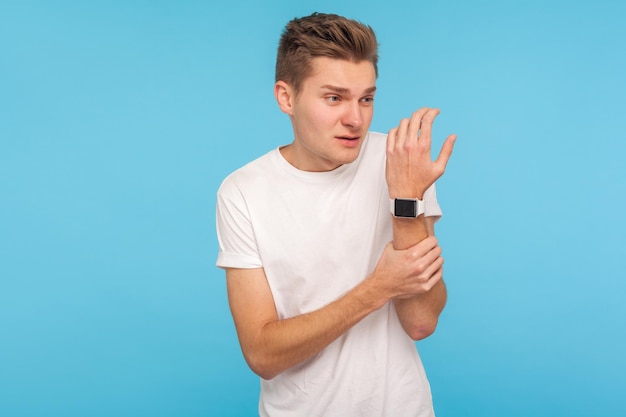 Injured hand. Man in white t-shirt holding arm and grimacing from pain, touching painful sprained wrist, suffering arthritis inflammation, pinched nerve. indoor studio shot isolated on blue background