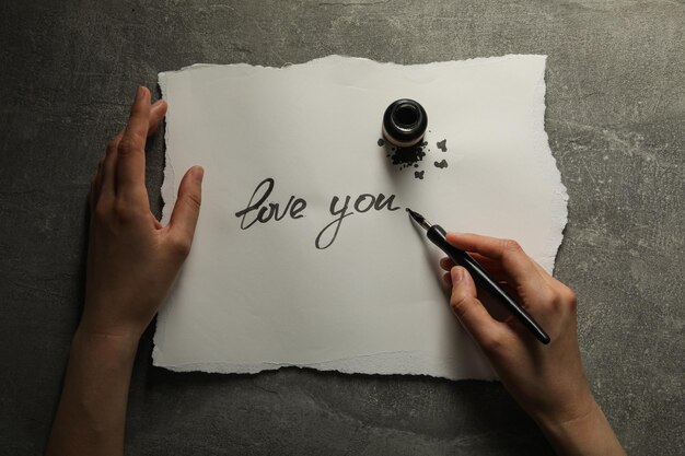 Ink bottle pen in hand and words Love you on paper on gray background top view