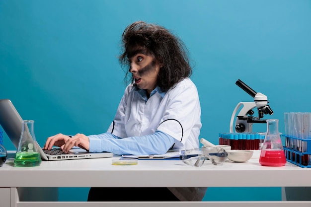 Insane angry biochemist with lunatic look after lab explosion sitting at desk and reviewing chemical experiment report. Wild foolish woman with smokey face researching discovery on laptop. Studio shot