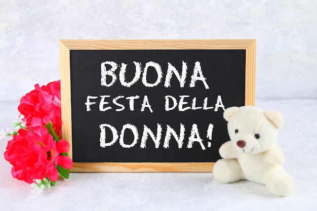 Photo the inscription on the chalkboard in italian: happy woman's day. pink flowers and teddy bear.