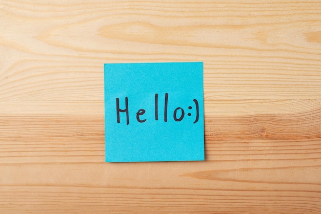 Inscription HELLO with a happy emojy on blue sticky note on a wooden background