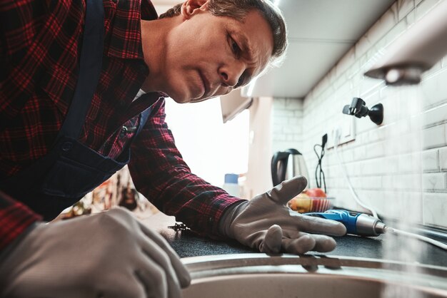 Photo inside view closeup of handsome plumber repairing sink in kitchen