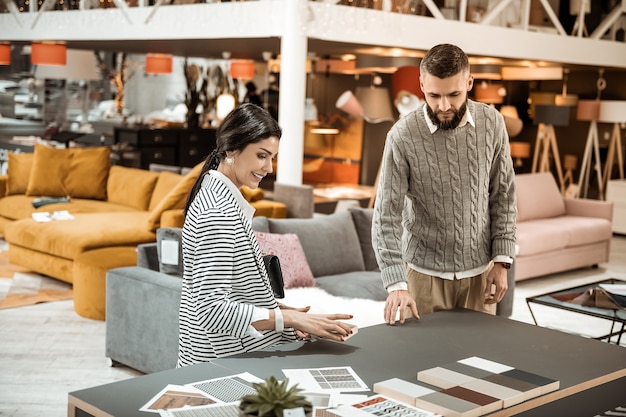 Inspecting colorful samples. Bearded focused man showing his wife wooden pieces and discussing color of table surface