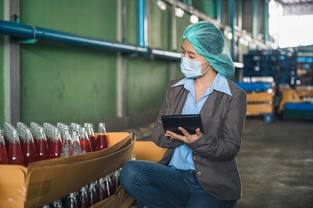 Inspector and supervisor asian woman checking with tablet a bottled fruit beverage on cardboard packaging in warehouse at beverage factory