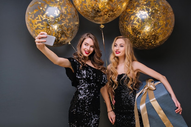 Inspired blonde woman holding big blue present and smiling at birthday party. Charming girl in black dress making selfie with friend in front of golden balloons.