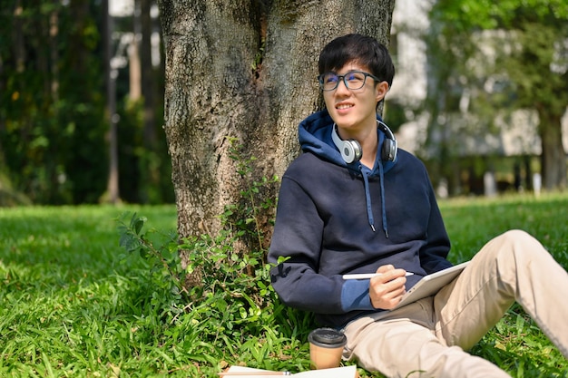 Inspired and happy young Asian male college student sitting under the tree in the park