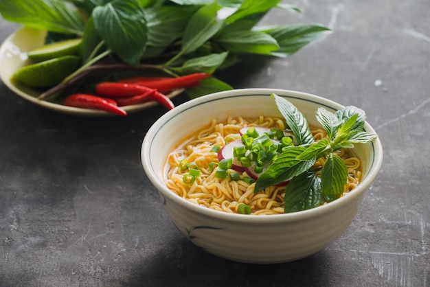 Instant noodles in bowl with fresh herbs garnish of cilantro and Asian basil lemon lime on dark stone background