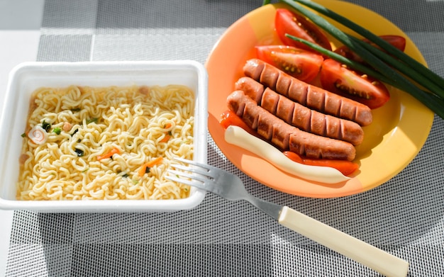 Instant noodles fried sausages green onions and tomatoes on table