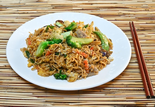 Instant noodles, stir-fried pork, vegetables, mushrooms, broccoli in white plate and wodden chopsticks.