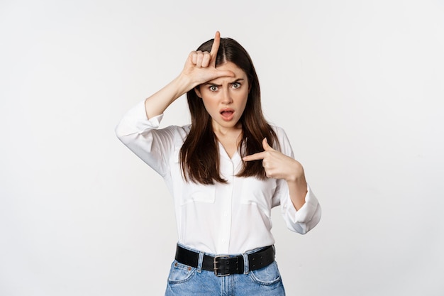 Insulted woman showing loser l word sign on forehead pointing at herself frustrated standing over wh...