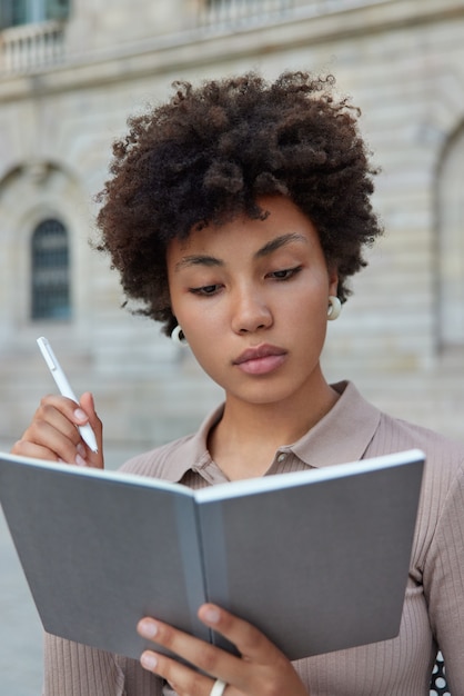 Intelligent female student with curly hair writes down information in notepad holds pen does homework or research for learning creats article has serious expression wears casual brown jumper