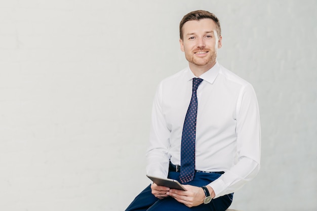 Intelligent lawyer in elegant clothes, holds digital tablet, spends free time in office, has pleasant smile on face