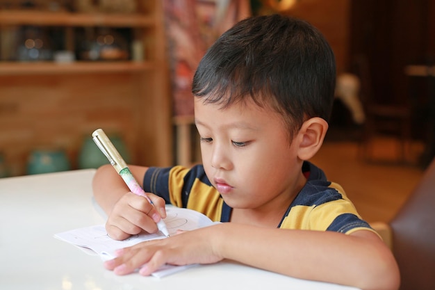 Intended Asian little boy drawing in a book with a pen on table in classroom