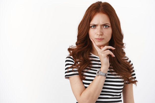 Intense serious-looking redhead girl express strong disbelief and doubts, furrow eyebrows, touch chin thoughtful, stare camera doubtful, pondering suspicious information