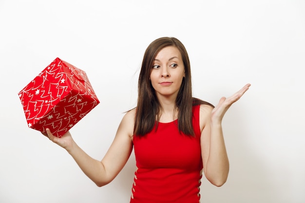 Interested and engaged young woman dressed in red dress and Christmas hat holds gift box, thinks that she was given on white background. Santa girl with present isolated. New Year holiday 2018 concept