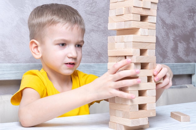 Interested happy little blond boy playing board game taking bricks from wooden tower keeping balance Board game Tower of wooden blocks Activity for strategy concentration agility