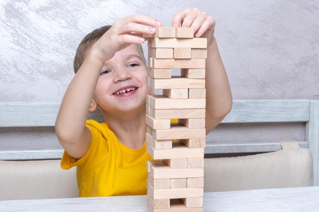 Interested happy little blond boy playing board game taking bricks from wooden tower keeping balance having fun