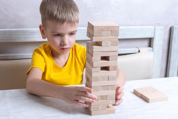 Interested happy little blond boy playing board game taking bricks from wooden tower keeping balance having fun