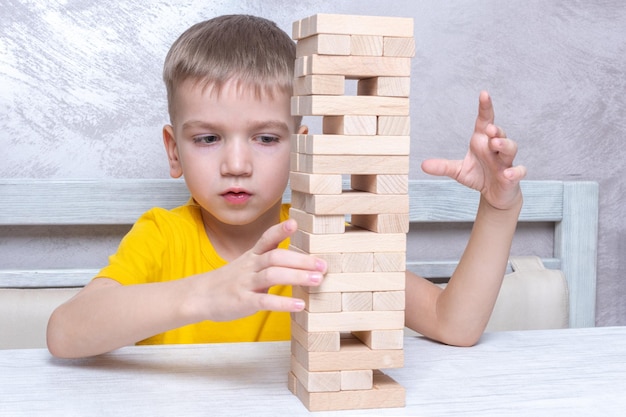 Interested happy little blond boy playing board game taking bricks from wooden tower keeping balance