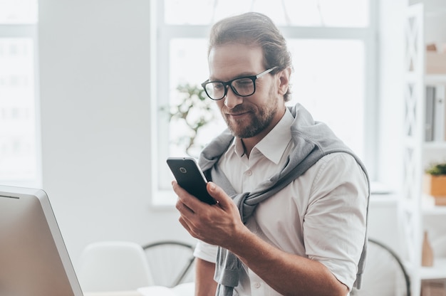 Interesting message. Handsome young man smiling and looking at the phone while sitting on the desk at his working place in creative office