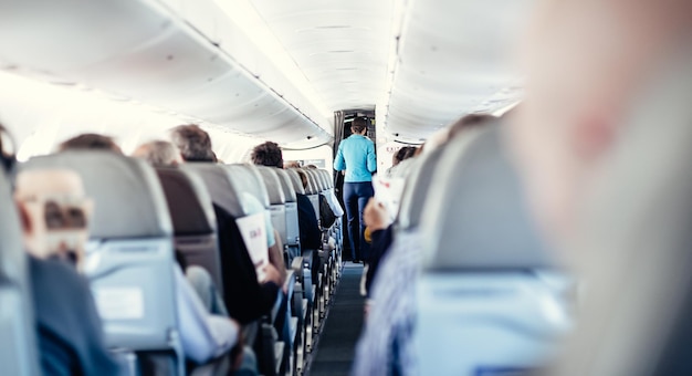 Interior of airplane with passengers on seats and stewardess in uniform walking the aisle serving pe