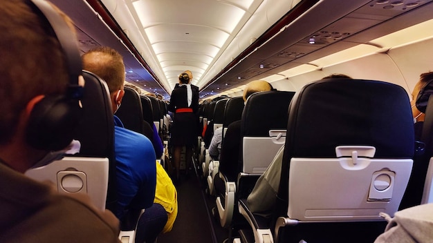 Interior of airplane with passengers on seats and stewardess in uniform walking the aisle