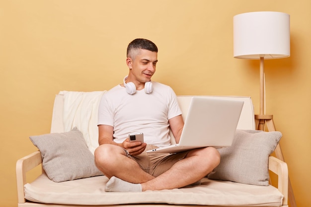 Internet browsing Home office Leisure surfing Positive attractive young man working on laptop computer and using smartphone sitting on comfortable couch at home