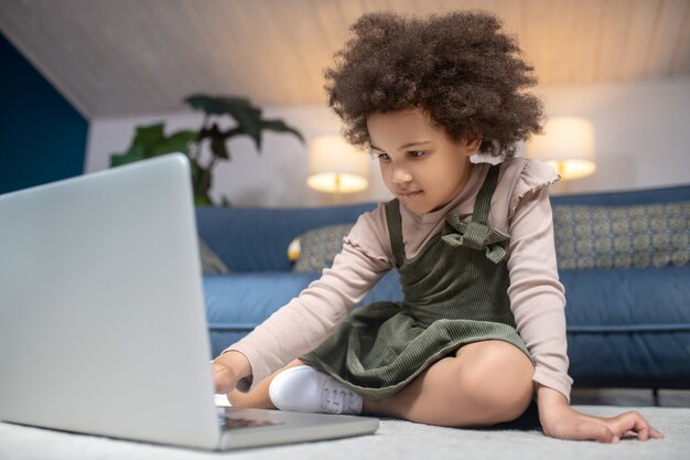 On internet. Little curly haired african american girl sitting near laptop on floor at home, focused interested