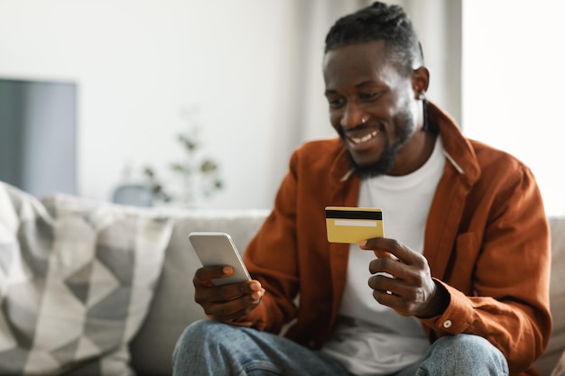 Internet mobile shopping concept Excited african american man using cellphone and holding credit card selective focus