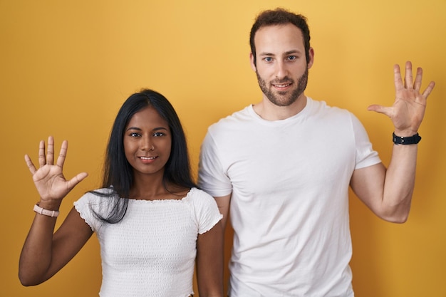 Interracial couple standing over yellow background showing and pointing up with fingers number ten while smiling confident and happy.