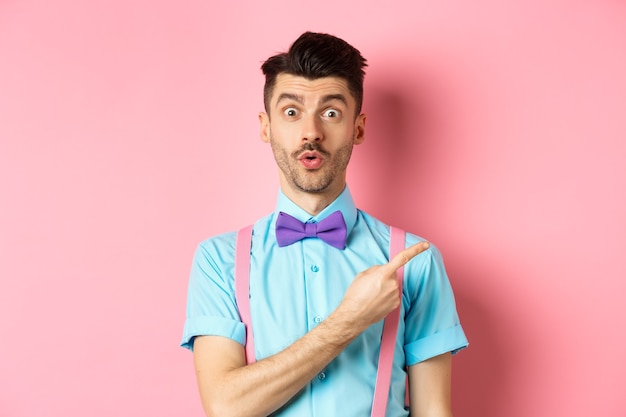 Intrigued and surprised cute guy with moustache showing awesome promo, looking curious and saying wow, pointing finger right at logo, standing on pink.