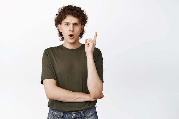 Intrigued young curly guy pointing finger up looking amazed at camera discussing promo above standing over white background