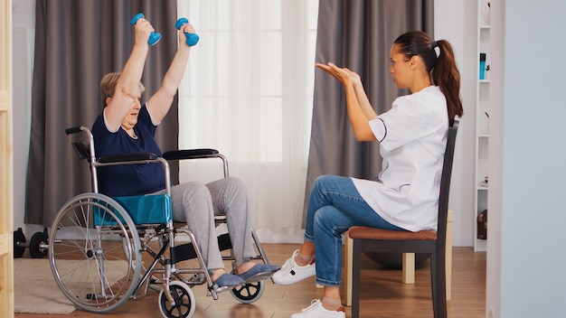 Invalid senior woman in wheelchair training with dumbbells during rehabilitation with nurse. Training, sport, recovery and lifting, old person retirement home, healthcare nursing, health support, soci