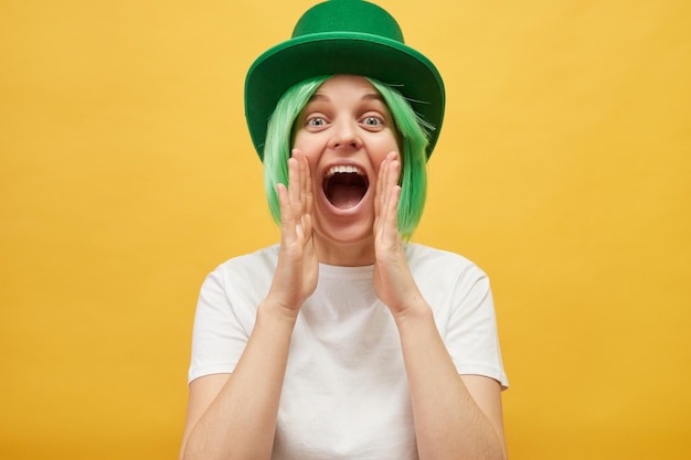 Inviting for St Patricks day party Excited Caucasian woman wearing green leprechaun hat standing isolated over yellow background screaming with hands near mouth making announce