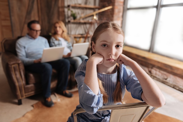 Involved depressed little kid waiting for parents care at home and feeling bored while father and mother working with laptops