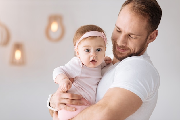 Involved in every moment. Happy joyful bearded father standing at home and hugging his little daughter while expressing tender emotions and positivity